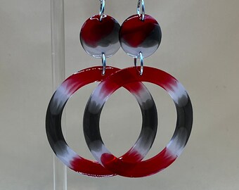 Puede incluir: Pendientes colgantes con un pequeño botón circular en rojo, negro y blanco conectado a un aro circular más grande en rojo, negro y blanco. Los pendientes están hechos de un material liso y brillante y cuelgan de ganchos de metal plateado.