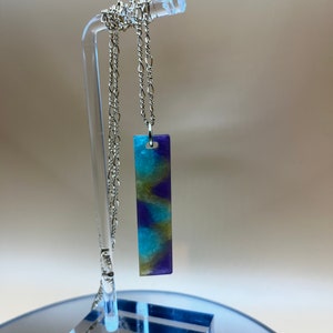 May include: A rectangular pendant necklace featuring a swirling resin design in shades of deep purple, bright turquoise, and metallic gold. The pendant hangs from a silver-toned metal chain with elongated oval links, suspended from a clear display stand.