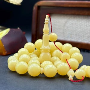 May include: Cream-colored prayer beads with a decorative central piece and a red string. The beads are round and smooth, and the central piece has an ornate design. The beads are likely made of a natural material.