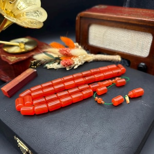 May include: A set of orange prayer beads, or tesbih, with a green string and decorative elements. The beads are rectangular and appear to be made of a translucent material. The beads are displayed on a black surface.