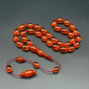 Puede incluir: Un juego de cuentas de oración de color ámbar anaranjado, también conocido como misbaha o tasbih, con cuentas ovaladas y una borla decorativa. Las cuentas están ensartadas en un cordón rojo, y el conjunto incluye una cuenta alargada decorativa.