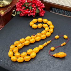 May include: A set of amber-coloured prayer beads, with oval beads and a decorative tassel. The beads are a warm yellow hue, with a smooth, polished finish. The tassel is attached with a red cord. The background is a dark surface.