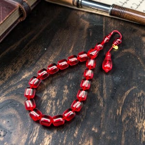 May include: A string of prayer beads featuring translucent, deep red, barrel-shaped beads. The beads are connected by a red cord, ending in a matching red tassel component and a small, circular gold-toned metal charm attached near the top.