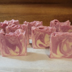 May include: Several rectangular bars of handmade soap arranged in a group. Each bar features a marbled pattern of soft pastel pink and creamy white swirls. The tops of the soap bars have a textured, uneven finish with visible peaks and ridges.