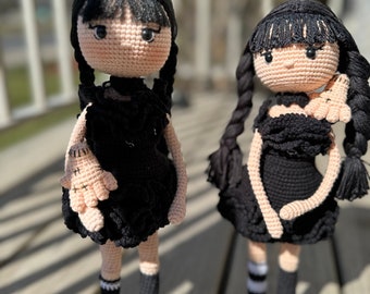 May include: Two crocheted dolls wearing black dresses with white and black striped socks and black shoes. The dolls have black hair and are holding crocheted dolls in their arms.