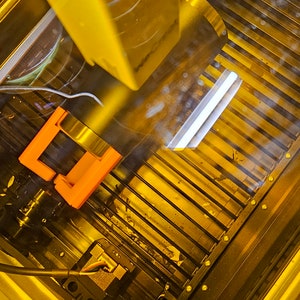May include: A close-up view of a 3D printer in operation. The printer is printing a small, orange object. The printer's nozzle is moving across a platform, and the object is being built up layer by layer.