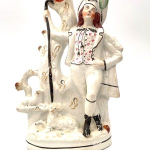 May include: A vintage porcelain figurine of a shepherd, holding a staff, with a sheep at his feet. The shepherd wears a white outfit with a floral vest and a hat with a pink and green plume. The figurine is painted in muted tones.