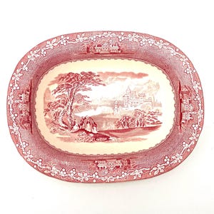 May include: Oval serving platter with a red and white toile design. The center features a landscape scene with a castle, trees, and figures. The border has a floral and architectural pattern. A classic piece for serving.