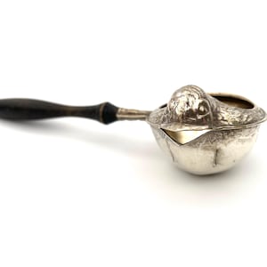 May include: A silver-toned metal tea strainer featuring a sculpted bird head design on the rim, a bowl-shaped mesh filter, and a long, slender dark brown wooden handle. The metallic surface shows intricate engraved details on the bird figure.