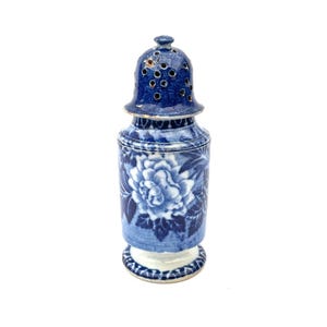 Op de afbeelding: Een blauw-witte keramische zoutstrooier met een bloemenontwerp. De shaker heeft een geperforeerd deksel en een gedetailleerd bloemmotief op de body. De basis is wit met een blauwe bloemenrand.