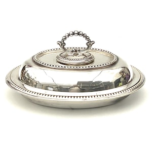 Puede incluir: Una fuente plateada con tapa. La fuente tiene forma redonda con borde de cuentas y asa en la tapa. La tapa también es redonda y tiene borde de cuentas. La fuente es reflectante y parece de metal.