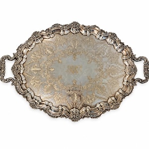 May include: An oval silver-toned serving tray featuring ornate, raised scrollwork along the scalloped edges and two integrated handles. The central surface displays an intricate, etched floral pattern with a weathered, metallic finish.
