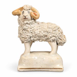 May include: A ceramic ram figurine featuring a textured, off-white woolly coat, smooth cream-colored legs, and a face with painted black eyes and nose. The ram has curved tan horns and stands on a rectangular, glazed off-white base.