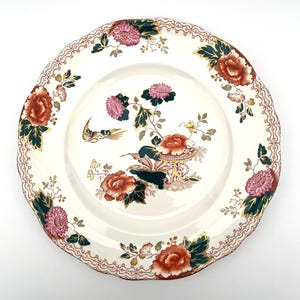 May include: A white ceramic plate with a scalloped edge, featuring a floral design in shades of red, pink, green, and gold. The plate has a detailed pattern of flowers and a bird, with a decorative border.