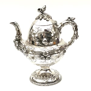 以下が含まれることがあります： 精巧な銀色のティーポット。花と葉の模様が施されています。ティーポットは、湾曲した注ぎ口、装飾的なハンドル、ドングリ型のつまみが付いた蓋が特徴です。ベースは、より多くの花のディテールを備えた台座です。