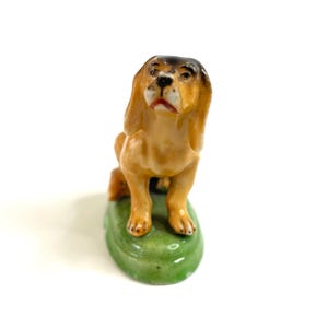 Könnte beinhalten: Keramikfigur eines Hundes mit braunem Körper und schwarzen Akzenten an Kopf und Ohren. Der Hund sitzt auf einem grünen, ovalen Sockel. Die Figur hat ein glänzendes Finish.