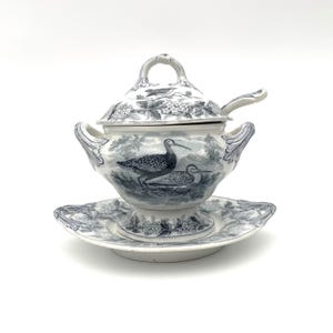 May include: A white and blue ceramic soup tureen with a lid, spoon, and matching saucer. The tureen features a bird and landscape design. The lid has a handle and the saucer has a scalloped edge. The tureen is a classic piece.