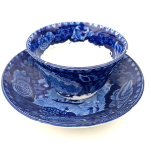May include: A blue and white ceramic teacup and saucer set featuring a detailed transferware pattern of a landscape with a boat, figures, and shell motifs. The interior of the cup is white, while the exterior and saucer display a deep blue.