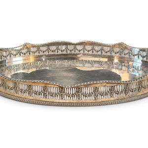 May include: An oval silver-toned serving tray featuring a decorative pierced gallery border with a repeating swag and tassel pattern. The tray has a polished reflective base and two integrated handles on the sides with a beaded edge.