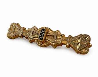 May include: A gold-toned metal bar brooch featuring intricate engraved star patterns on the flared ends. The center displays a circular element with a rectangular black enamel inlay decorated with small gold cross motifs and geometric detailing.