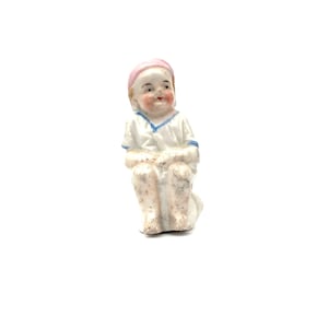 May include: A small, antique porcelain figurine of a seated child. The child wears a white outfit with blue trim and a pink cap. The figurine has rosy cheeks and a detailed, hand-painted design.