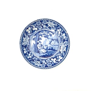 Peut inclure: Assiette décorative ronde bleue et blanche. Elle présente une scène centrale d'une personne et d'un mouton dans un paysage, entourée d'une bordure florale. Le bord a un motif floral répétitif.