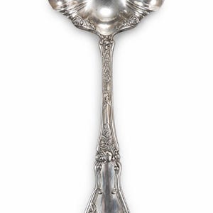 May include: A silver-toned metal ladle featuring an ornate, scalloped bowl and a long handle decorated with embossed floral motifs, including rose blossoms and leaves, extending from the base of the bowl down to the rounded handle end.