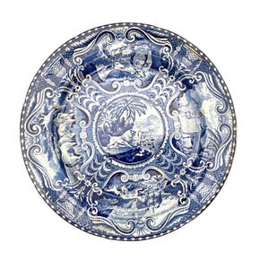 Puede incluir: Un plato decorativo redondo, azul y blanco, con un diseño paisajístico detallado. El plato presenta una escena central con un león y palmeras, rodeada de adornos ornamentados y viñetas paisajísticas adicionales. El borde tiene un borde estampado.