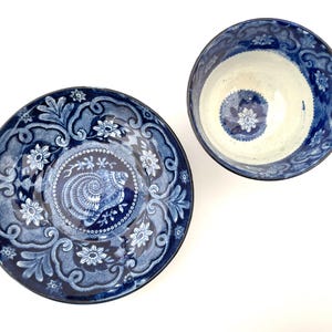 May include: A ceramic tea cup and matching saucer set featuring a deep blue and white color palette. The saucer displays a central seashell motif surrounded by intricate floral and scroll patterns. The cup interior is cream with a blue floral design.