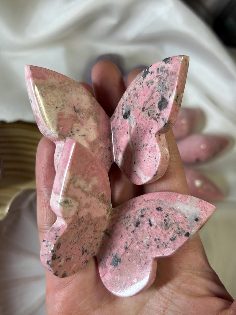 May include: Pink and gray butterfly-shaped stone carvings. The carvings have a polished finish and are held in a hand. The stone has a mix of pink, gray, and black colors.
