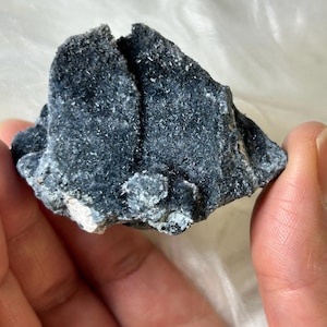 May include: A dark blue mineral specimen with a rough, textured surface. The rock has a crystalline structure and is held in a hand against a soft, white background. The mineral appears to be a cluster of small, needle-like crystals.