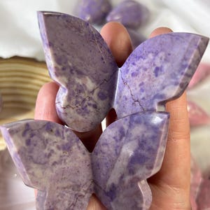 May include: Two carved butterfly figurines in shades of purple and white are held in a hand. The butterfly wings have a textured appearance with natural veining. Other butterfly figurines are visible in the background.