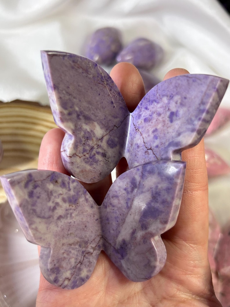 May include: Two carved butterfly figurines in shades of purple and white are held in a hand. The butterfly wings have a textured appearance with natural veining. Other butterfly figurines are visible in the background.