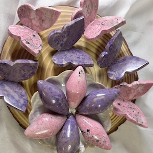 May include: A collection of pink and purple butterfly-shaped stones arranged on a wooden slice and a decorative dish. The stones have a marbled appearance with dark speckles. The arrangement resembles a flower with butterfly wings around it.