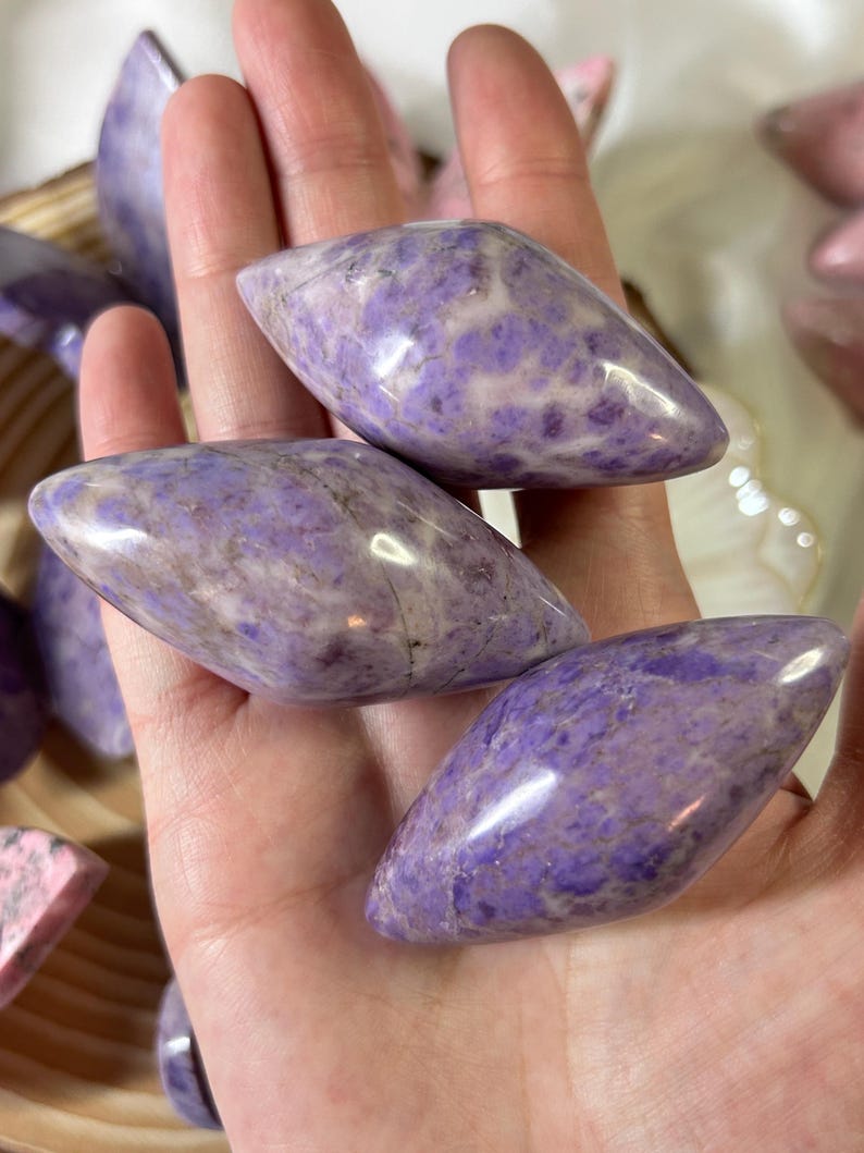 May include: Close-up of several polished, oval-shaped gemstones. The stones are a mix of lavender and white, with a smooth, glossy finish. The gemstones are held in a hand, with other stones visible in the background. The stones are a beautiful shade of purple.