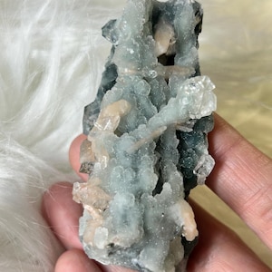 May include: A natural mineral specimen featuring a cluster of light blue and peach-colored crystals. The crystals have a textured, bubbly appearance, with some areas appearing translucent. The mineral sits against a soft, white background.