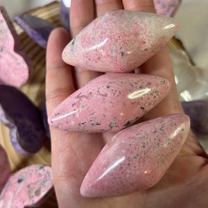 May include: Three polished pink stones with black speckles are held in a hand. The stones have a smooth, glossy surface and a rounded, elongated shape. Other butterfly-shaped stones in shades of pink and purple are visible in the background.