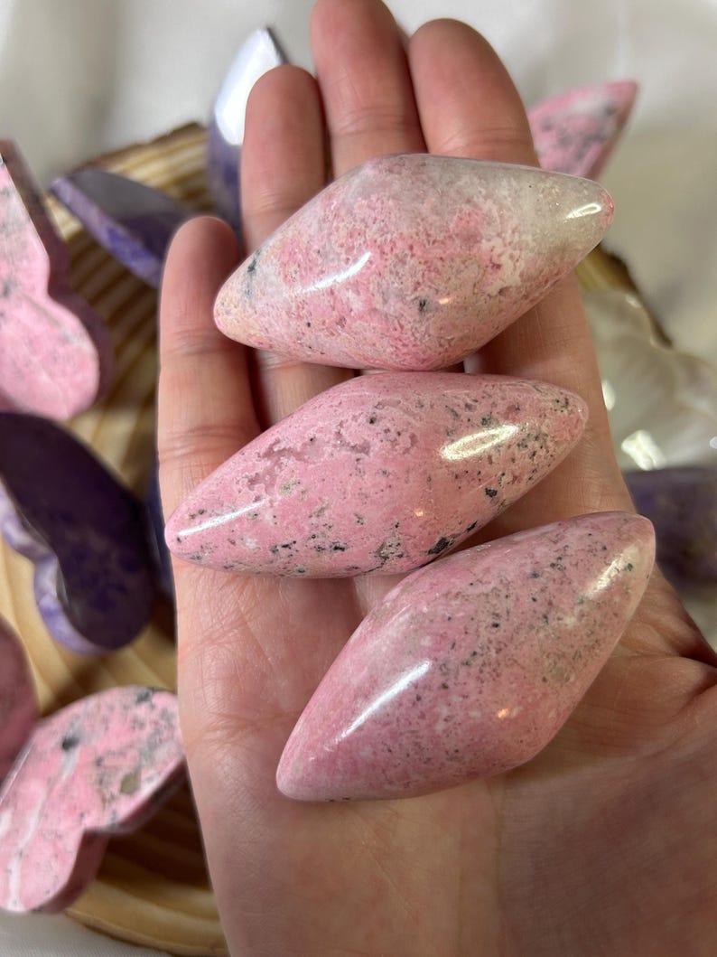 May include: Three polished pink stones with black speckles are held in a hand. The stones have a smooth, glossy surface and a rounded, elongated shape. Other butterfly-shaped stones in shades of pink and purple are visible in the background.