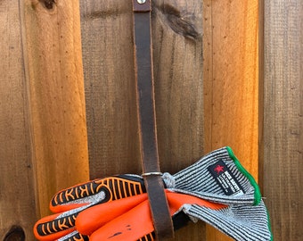 Puede incluir: Una correa de cuero marrón con un gancho plateado cuelga de una valla de madera. Un par de guantes de trabajo naranja y gris están unidos a la correa.