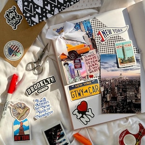 May include: A scrapbook featuring New York City imagery, including a yellow taxi, the Empire State Building, and the Statue of Liberty. Stickers with text like "I Love NY" and "It will always be New York or nowhere" are arranged alongside scissors, markers, and tape.