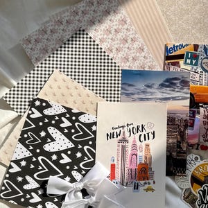 May include: A collection of stationery items including patterned papers with floral, gingham, and heart designs, a postcard featuring a New York City skyline illustration with the text Greetings from NEW YORK CITY, and white ribbon bows.