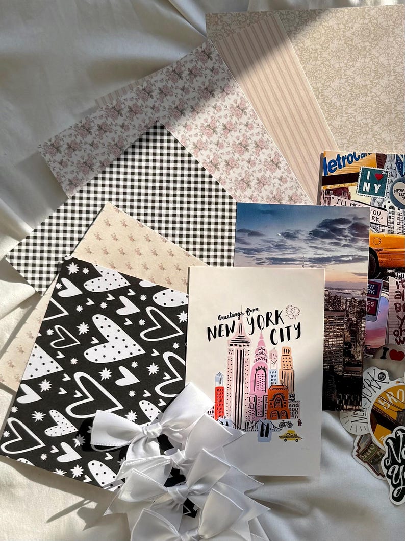 May include: A collection of stationery items including patterned papers with floral, gingham, and heart designs, a postcard featuring a New York City skyline illustration with the text Greetings from NEW YORK CITY, and white ribbon bows.