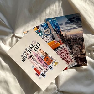 May include: Three rectangular postcards featuring New York City themes. One postcard displays a stylized illustration of skyscrapers with the text Greetings from NEW YORK CITY. Another shows a yellow taxi and city skyline. The third is a night view.