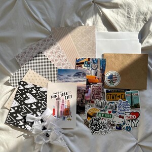 May include: A collection of stationery items including patterned paper sheets, white envelopes, a brown envelope, and a stack of New York City themed postcards and stickers. Three white ribbon bows are attached to a black and white heart-patterned card.