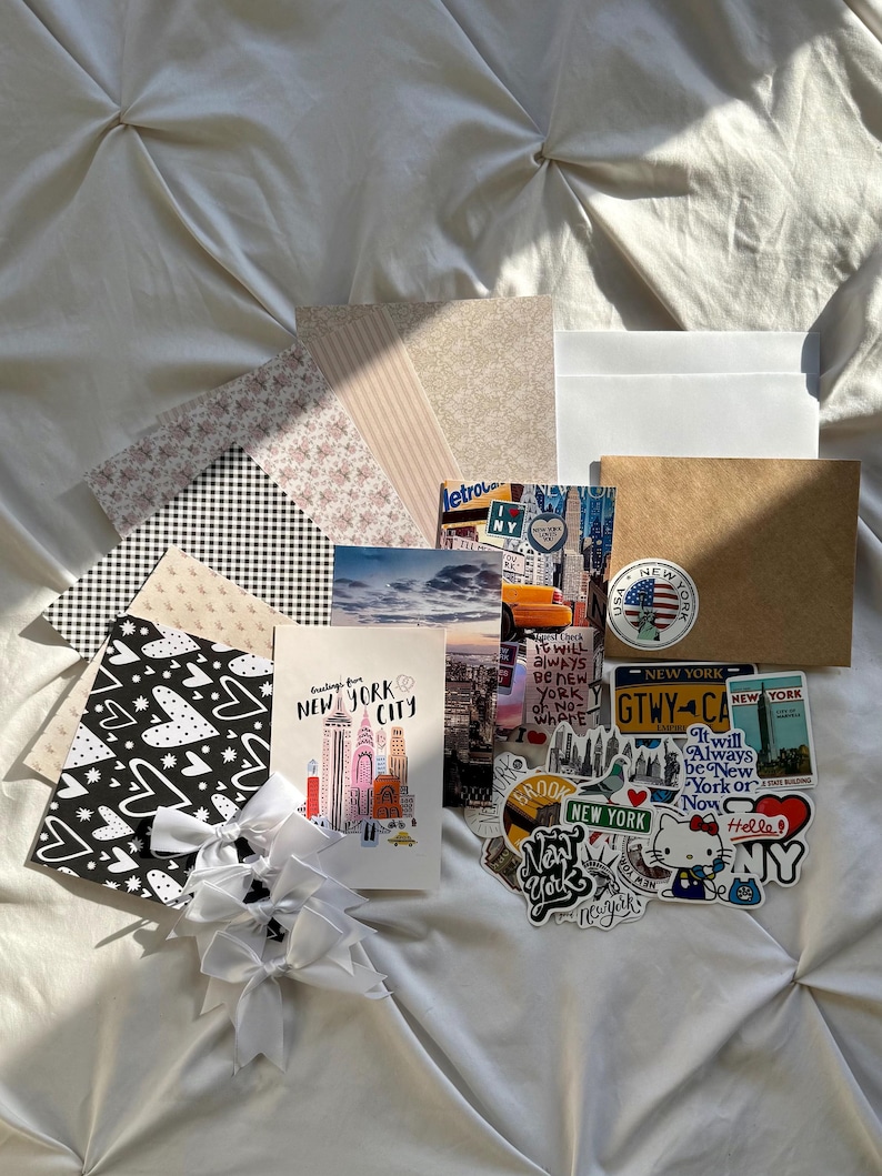 May include: A collection of stationery items including patterned paper sheets, white envelopes, a brown envelope, and a stack of New York City themed postcards and stickers. Three white ribbon bows are attached to a black and white heart-patterned card.