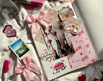 May include: A flat lay of wedding-themed stationery and decorations. Includes pink envelopes, a notebook, stickers, a pink bow, scissors, and a pink Sharpie marker. The stickers feature wedding rings, a car, and the words "Our Wedding Day."