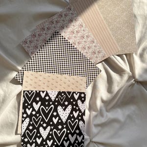 May include: A collection of square paper sheets featuring various patterns including a black background with white hearts and stars, a black and white gingham check, a floral print, vertical beige stripes, and a beige damask-style design.