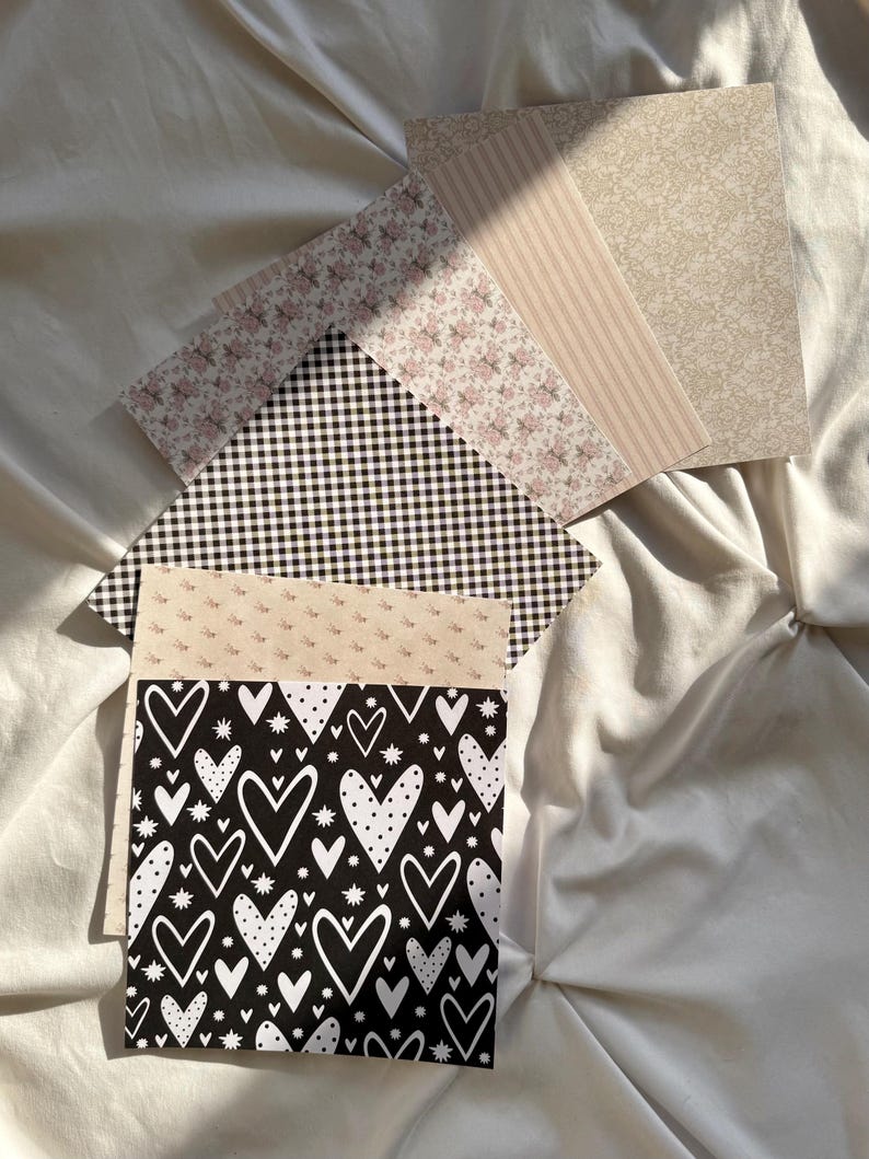 May include: A collection of square paper sheets featuring various patterns including a black background with white hearts and stars, a black and white gingham check, a floral print, vertical beige stripes, and a beige damask-style design.