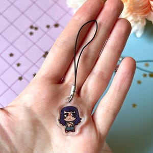 May include: A clear acrylic phone charm with a cartoon illustration of a girl with long dark blue hair, wearing a blue and white shirt and blue pants. The charm is attached to a black string.