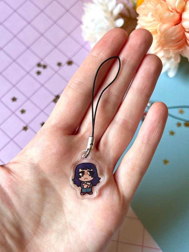 May include: A clear acrylic phone charm with a cartoon illustration of a girl with long dark blue hair, wearing a blue and white shirt and blue pants. The charm is attached to a black string.